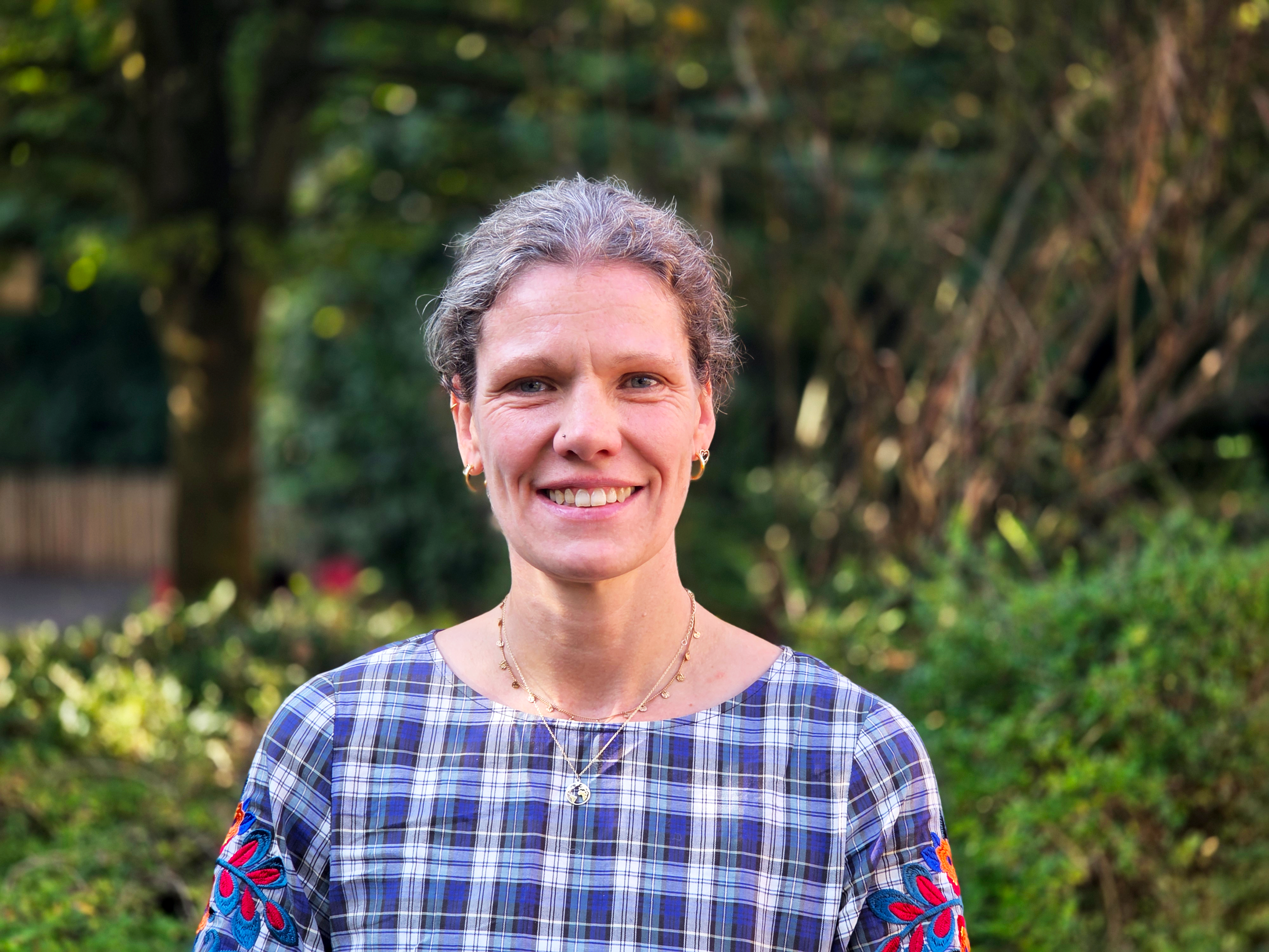 Auf dem Bild ist eine lächelnde Frau als Porträt zu sehen, vor einem unscharfen grünen Hintergrund im Freien. Sie trägt ein blau-weiß kariertes Oberteil und Schmuck an den Ohren und am Hals.