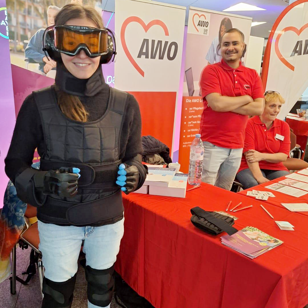 Auf dem Foto sind drei Personen an einem Stand der AWO (Arbeiterwohlfahrt) zu sehen. Im Hintergrund sind Banner mit dem AWO-Logo zu erkennen, was darauf hinweist, dass es sich um eine Veranstaltung oder Messe der Organisation handelt. Die Person im Vordergrund trägt eine schwarze Weste und Handschuhe mit blauen Fingerspitzen, was auf eine virtuelle Realität oder ein physisches Training hinweisen könnte. Auf dem Tisch vor ihnen liegen Broschüren und ein Tablet. Die Personen im Hintergrund tragen ein rotes Shirt mit dem AWO-Logo, was darauf hindeutet, dass sie Mitarbeiter der Organisation ist.