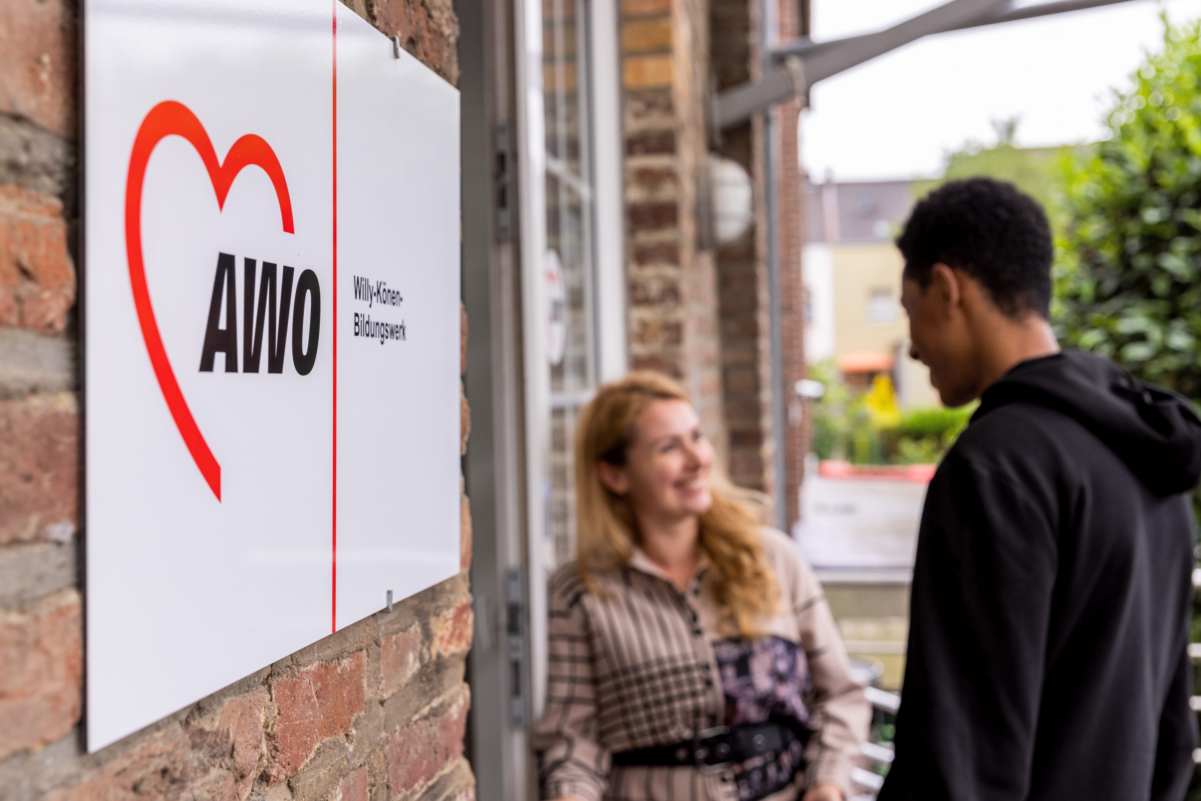 Auf dem Foto ist ein Schild mit dem Logo der AWO (Arbeiterwohlfahrt) und dem Zusatz "Willy-Könen-Bildungswerk" zu sehen, das an einer Backsteinwand angebracht ist. Im Hintergrund unterhalten sich zwei Personen freundlich vor dem Gebäude – eine Frau mit langen blonden Haaren und ein junger Mann mit dunklen Haaren im schwarzen Hoodie.