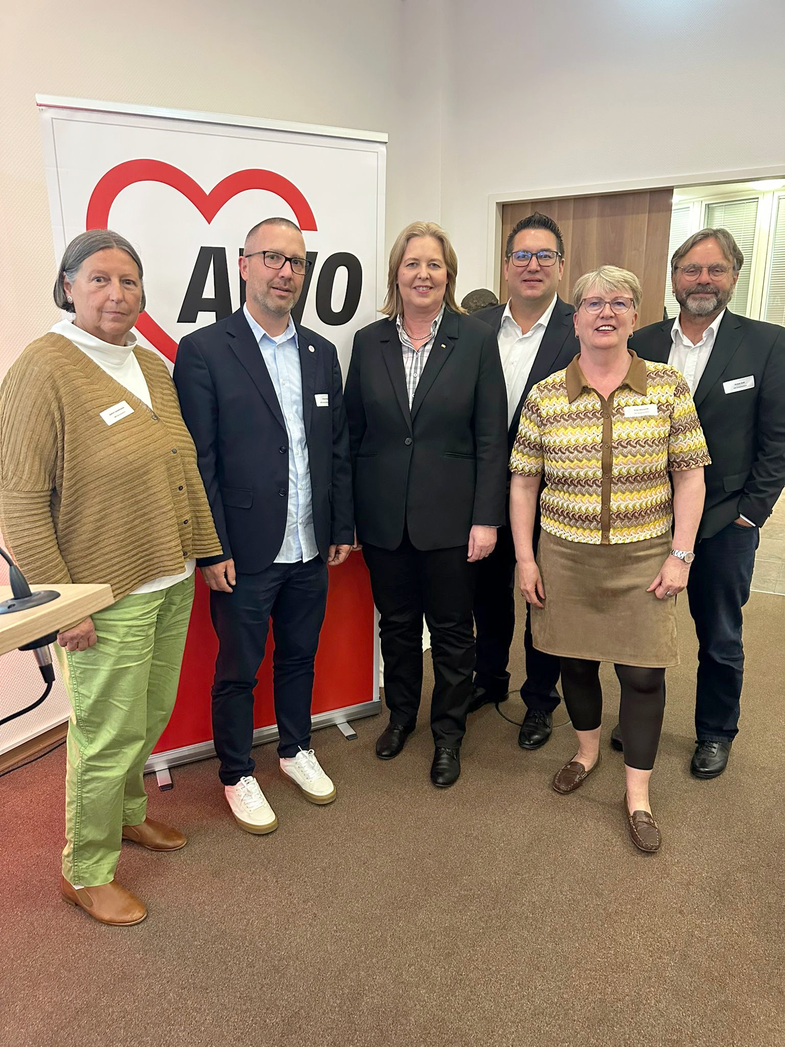 Auf dem Foto sind sechs Personen zu sehen, die gemeinsam vor einem Roll-Up mit dem Logo der AWO (Arbeiterwohlfahrt) stehen. Das AWO-Logo zeigt ein rotes Herz mit dem Schriftzug „AWO“ darin. Die Personen wirken formell oder semi-formell gekleidet. Die Personen tragen Namensschilder, was weiter auf einen offiziellen oder öffentlichen Anlass hindeutet. Der Hintergrund zeigt einen Konferenzraum oder Bürobereich.